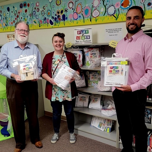 Three people standing in front a book shelf full of toys in plastic bags with a sign on top of the shelf reading "Play2Learn"